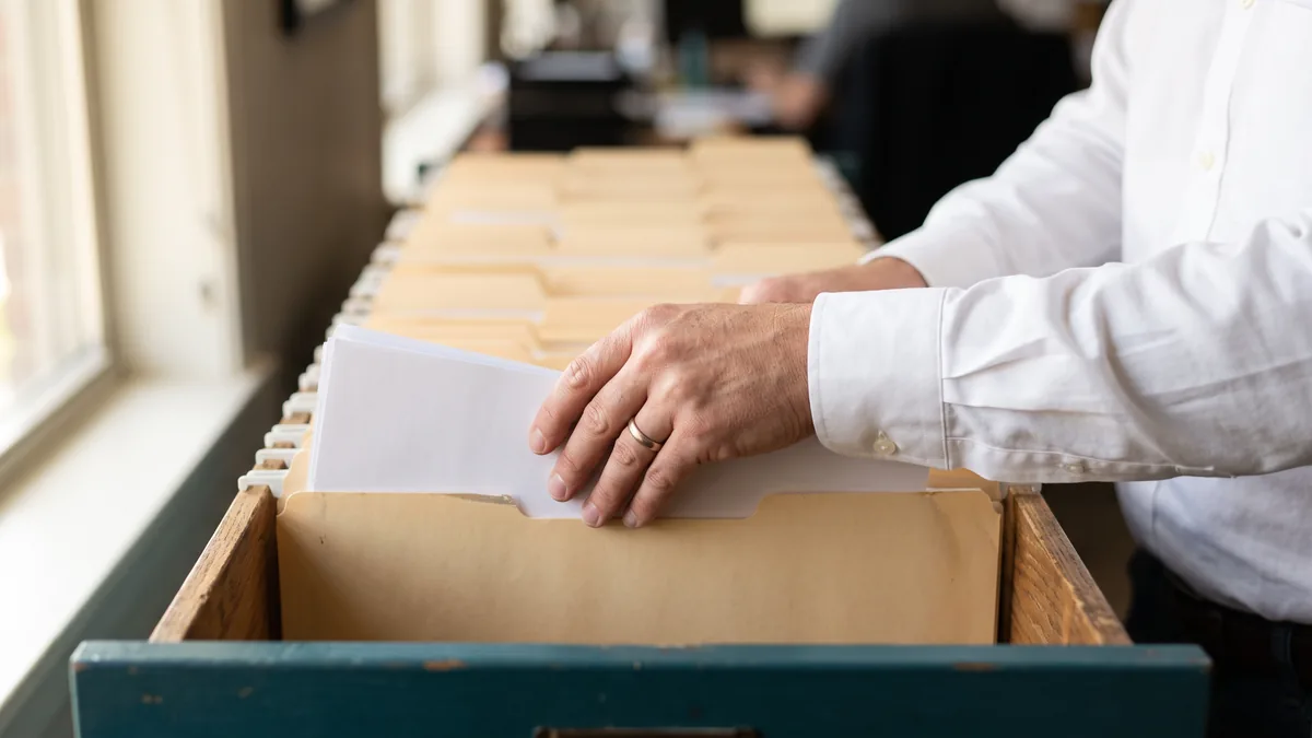 Hands lowering a sheaf of pages into an open manila folder in a wooden filing drawer