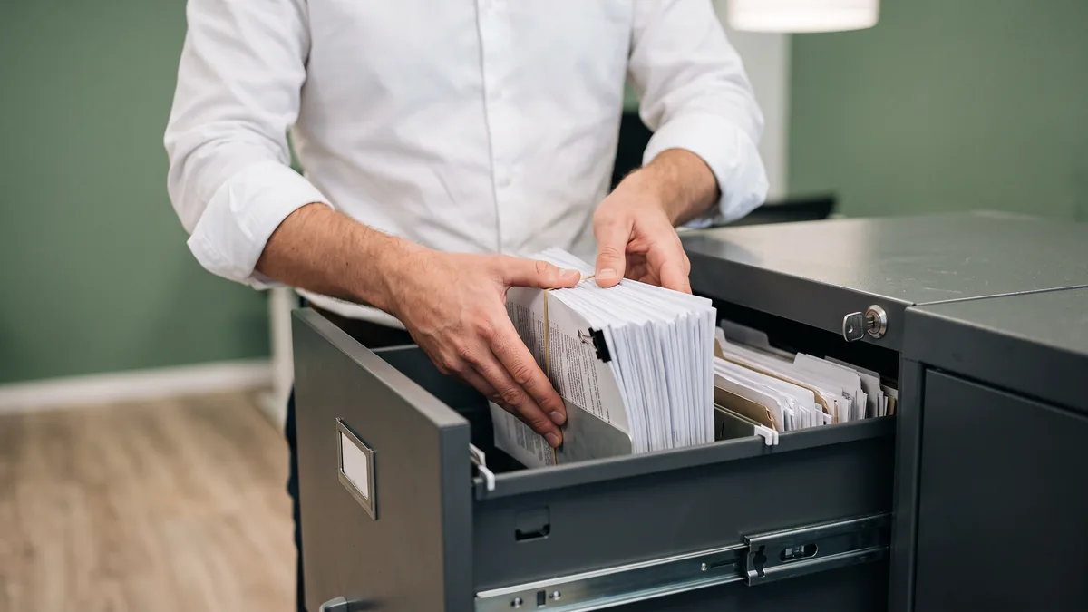 Hands placing a bundle of printed client records into a locking steel document cabinet