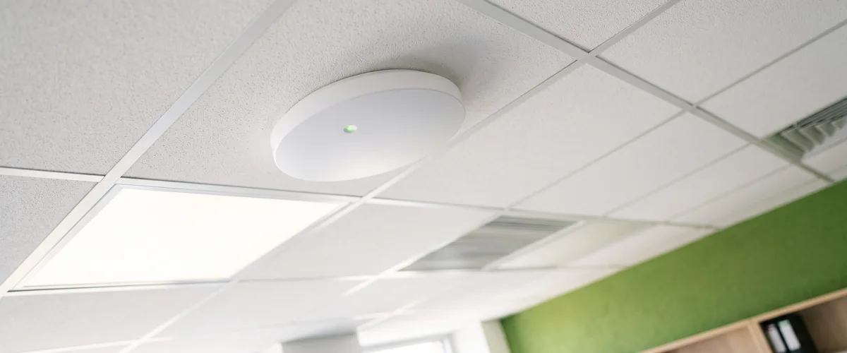 White ceiling-mounted business Wi-Fi access point installed on a drop-ceiling tile in a bright small office