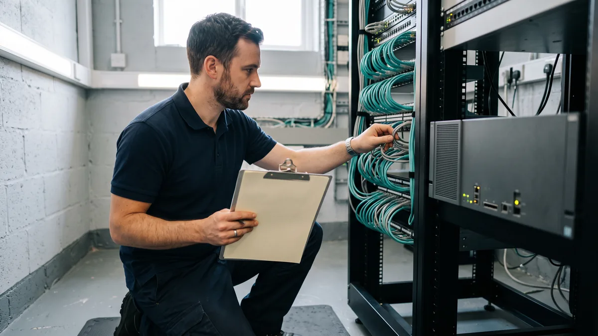 IT technician with a clipboard inventorying a small business file server and network rack before a Microsoft 365 migration